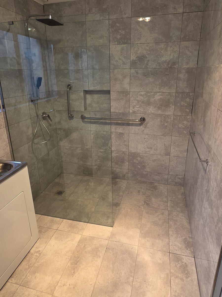 Walk-in shower with hobless level entry, frameless glass screen, vertical and horizontal grab rails, recessed niche and a floor waste set into the tiled shower base