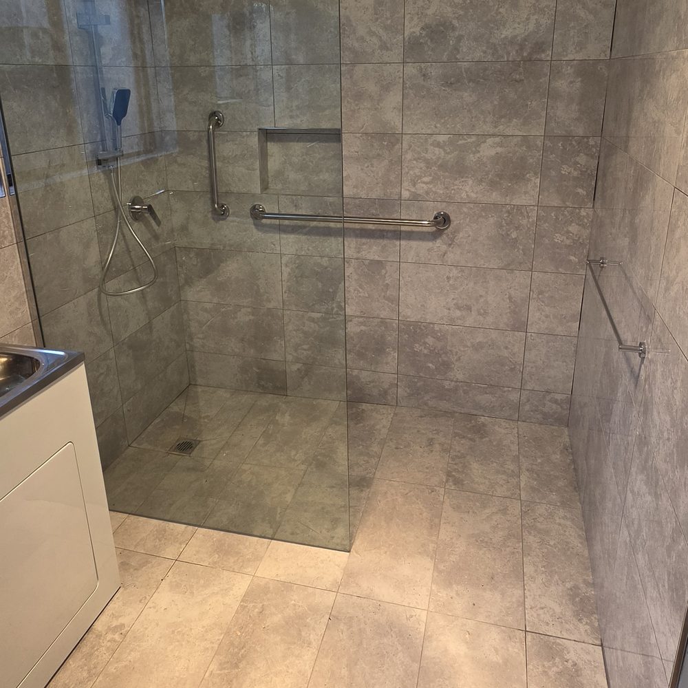 Completed NDIS bathroom modification at Blackshaws Road, Altona: level-access walk-in shower with frameless glass screen, vertical and horizontal grab rails and a floor waste set into the tiled base